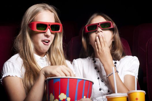 two girls look three-dimensional cinema, sitting in the glasses, eat popcorn, drink drink