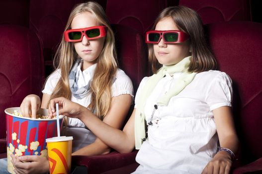 two girls look three-dimensional cinema, sitting in the glasses, eat popcorn, drink drink