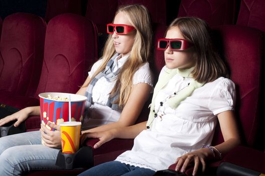 two girls look three-dimensional cinema, sitting in the glasses, eat popcorn, drink drink