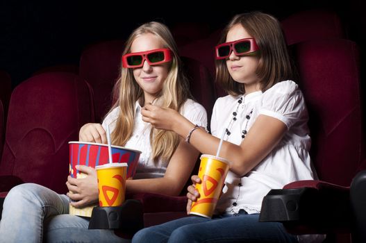 two girls look three-dimensional cinema, sitting in the glasses, eat popcorn, drink drink