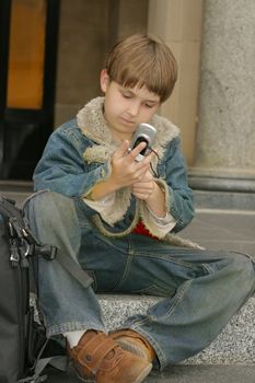 Schoolboy in jeans and a jacket sitting on steps with a cellphone