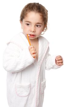 A small preschool age child brushing her teeth with an orange coloured toothbrush.  She is wearing a white bathrobe edged with pink.