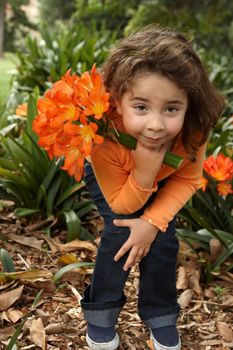 Little girl holding a bunch (umbels) of lilies  (clivia miniata).  She is wearing navy jeans and an orange twin set and  standing in the garden bed.