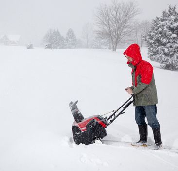 Senior man in red coat using a snow blower during a blizzard on home drive