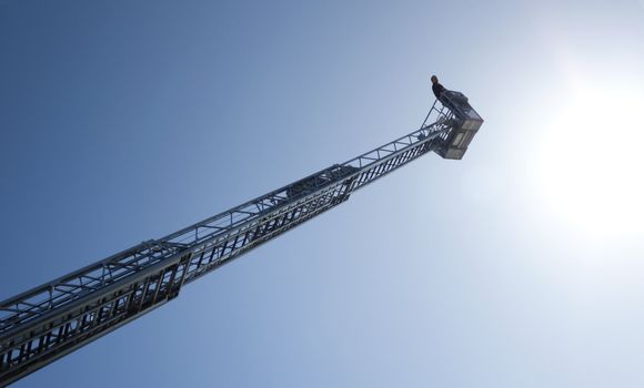 Male person in a basket on top of a high ladder, against a sunny blue sky