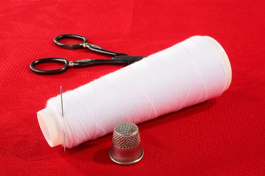 The coil with white threads and a needle on a red background, nearby a thimble for sewing.