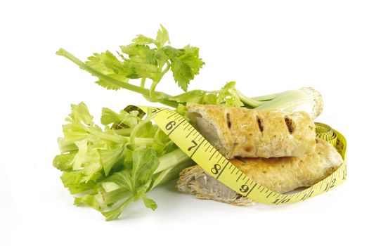 Contradiction between healthy food and junk food using celery and sausage roll with a tape measure on a reflective white background 