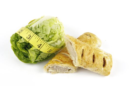 Contradiction between healthy food and junk food using a green salad lettace and sausage roll with a yellow tape measure on a reflective white background 