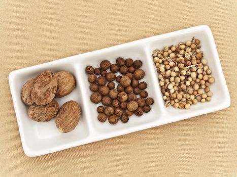 Coriander, black pepper seeds and nutmeg in flavouring plate at beige textile