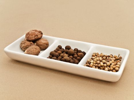 Coriander, black pepper seeds and nutmeg in flavouring plate at beige textile