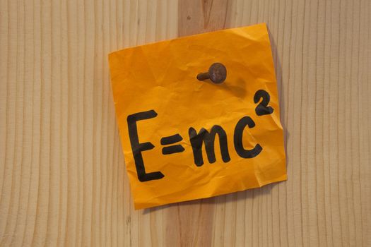 Albert Einstein well known physical formula describing equivalence of matter mass and energy, handwritten on bright orange reminder note nailed to wooden wall.