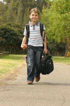 Boy walking to school.  He has a backpack, carrying a small portable computer and is holding an apple in his hand
