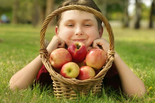 A child with a basketful of Pink Lady apples.  He is smiling and laying in the grass.