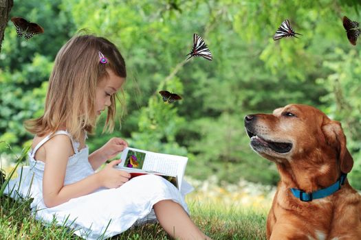 Little girl sits under a tree reading a book about butterflies as her faithful dog sits nearby watching butterflies fly around them.