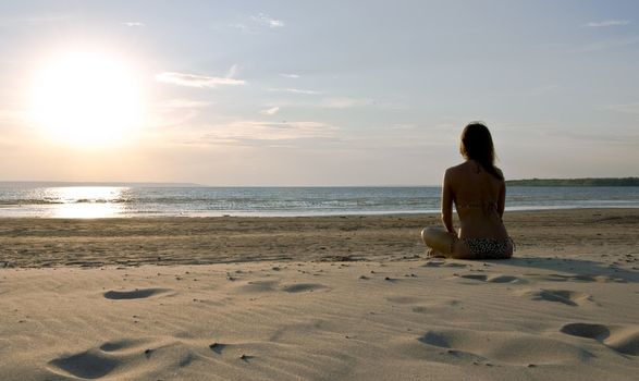 Young girl sitting on a deserted beach back to the camera and admires the sunset. Landscape.