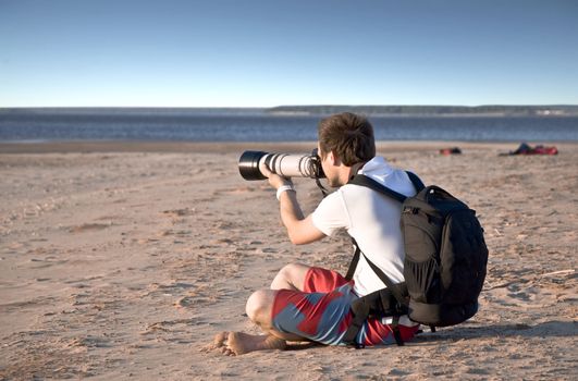 Photographer with long lens pictures of water views, sitting on a sandy beach. Summer landscape against the blue sky.