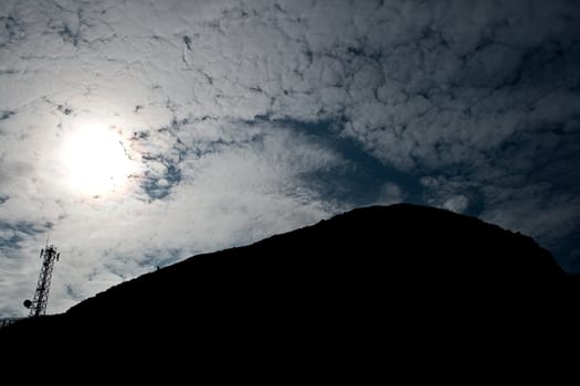 Hill silhouette with communications tower, sun with trail in the clouds.