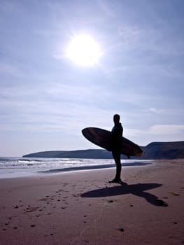 Silhouette of a surfer with surfboard standing on a beach with the sun above.