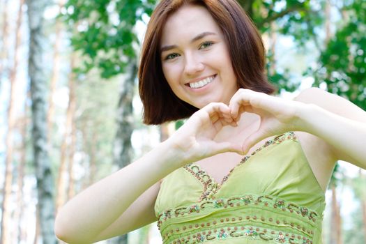 Close-up portrait of a young smiling gilr with a heart sign at the park