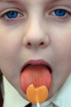 young blue eyed girl licking an orange lollipop into a heart shape