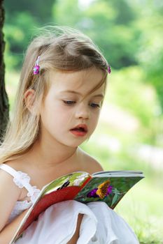 Little girl sits outdoors under a tree reading a book about butterflies.