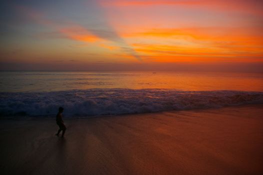 Boy on a beach at sunset with waves rolling towards him. Dreamland beach, Bali, Indonesia.