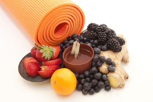 Bright orange yoga mat with ginger tea in a distinctive cup surrounded by bright berries, ginger root, and citrus.  On white background.  