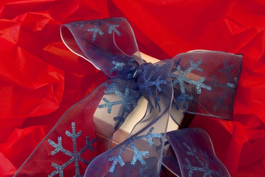 Vibrant holiday image of white jewelry box wrapped in a blue, snowflake accented ribbon against a background of red tissue paper.