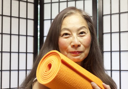 Mature woman with bright yoga mat portrays excitement at fitness and health. Asian woman and shoji screen background depict fresh, alternative nature of yoga with today's medicine.