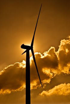 Wind turbine in the back light of the sun with yellow sky and clouds.
