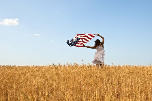 Beautiful young girl holding an American flag in the wind in a field of rye. Summer landscape against the blue sky. Horizontal orientation.