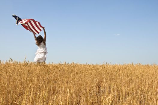 Beautiful young girl holding an American flag in the wind in a field of rye. Summer landscape against the blue sky. Horizontal orientation.