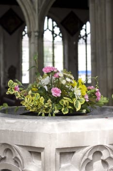 A church christening font with an inset floral decoration of green and yellow foliage and pink carnation flowers. The arched pillars of the church visible in soft focus to the background. Location in Salisbury Wiltshire, UK england.