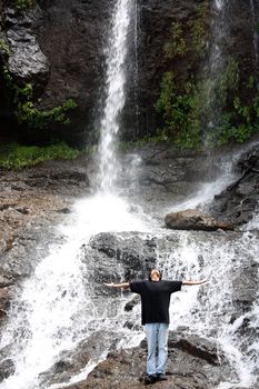 A young Indian man standing at the bottom of a tall waterfall enjoying the bliss in nature.
