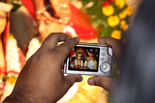 Two hands holding a small digital camera, clicking the traditional Indian wedding couple.