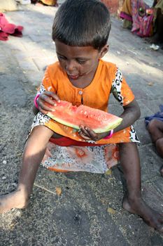A hungry beggar girl from India eating a watermelon, sitting on a street.