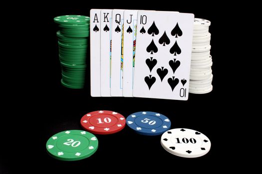 A poker hand of a royal flush of spades leaning against a stack of gambling chips, on black studio background.