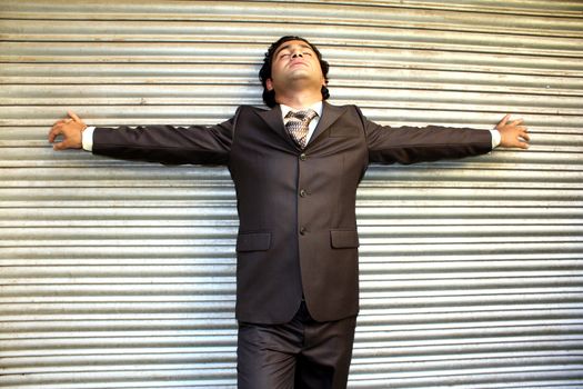 A stressed Indian businessman leaning on a shutter with his arms stretched.