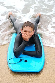 Smiling teenage surfer in a wetsuit laying on his bodyboard on the beach.
