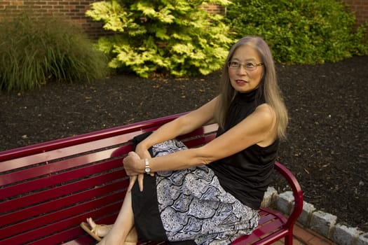 Mature, senior Asian woman seated on a red metal bench has feet up to take a break