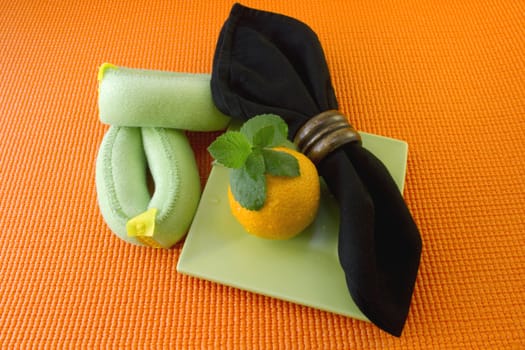 Green wrist weights, an orange, mint leaves, and a black napkin agains an orange yoga mate reflect importnat points in a healthy lifestyle