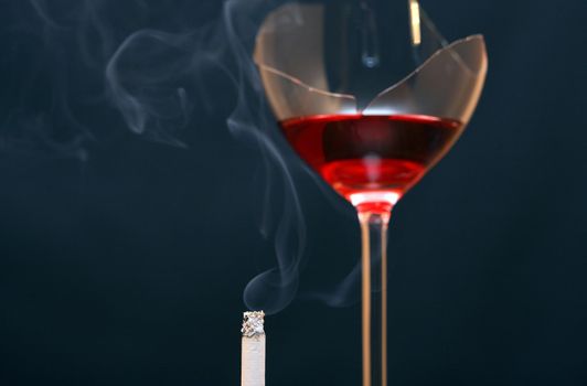 Smoking cigarette on dark background with broken goblet of red wine
