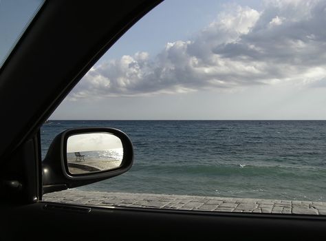 Seascape viewed from car with bench reflection in car mirror.