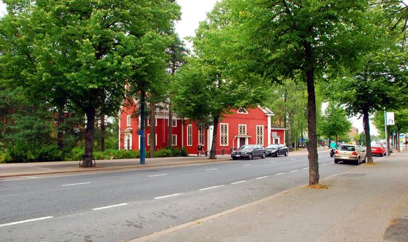 One of the main streets of Hyvinkaa, a city not far from Helsinki, we can see cars and on the other side the red church of Hyvinkaa, with many trees all around.