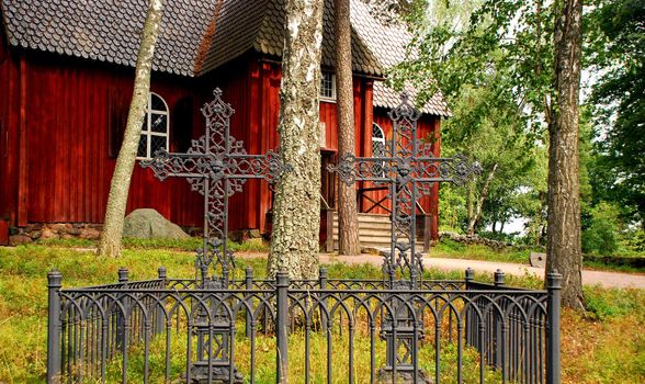 We can see two crosses made of metal, in frot of a finish red church, rounded by nature, many trees. This photograph was taken in a museum of finish life, in the island named suomenlinna, in helsinki.