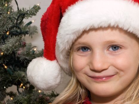 Young girl wearing a santa hat with christmas tree behind her