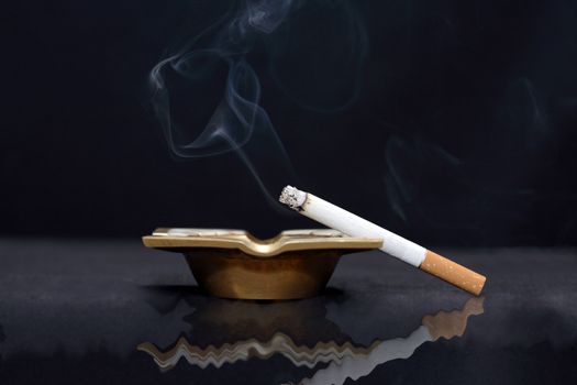 Closeup of smoking cigarette lying on old bronze ash-tray on dark background