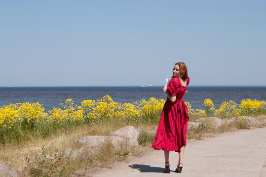 Young lady near the sea in long red dress. Near beauty yellow flowers