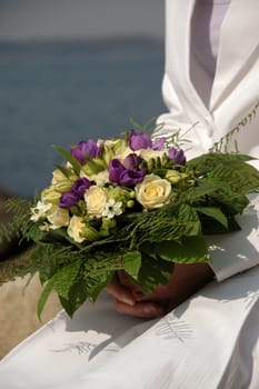 Bride is sitting by the ocean, holding a bouquet of flowers.