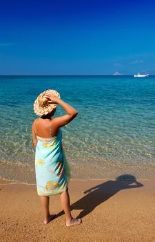 Young woman in a pareo and straw hat, standing on a Mediterranean sandy beach and looking into the blue sea